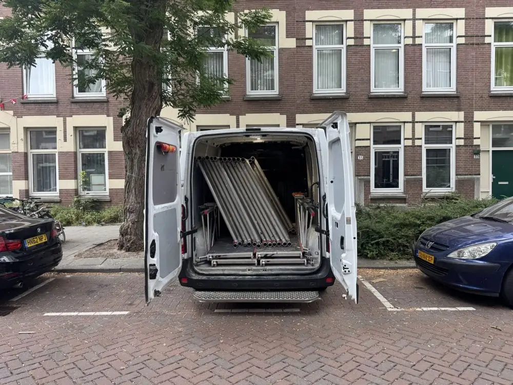 Bezorgbus met geopende achterdeuren vol steigeronderdelen, geparkeerd voor een rij woningen